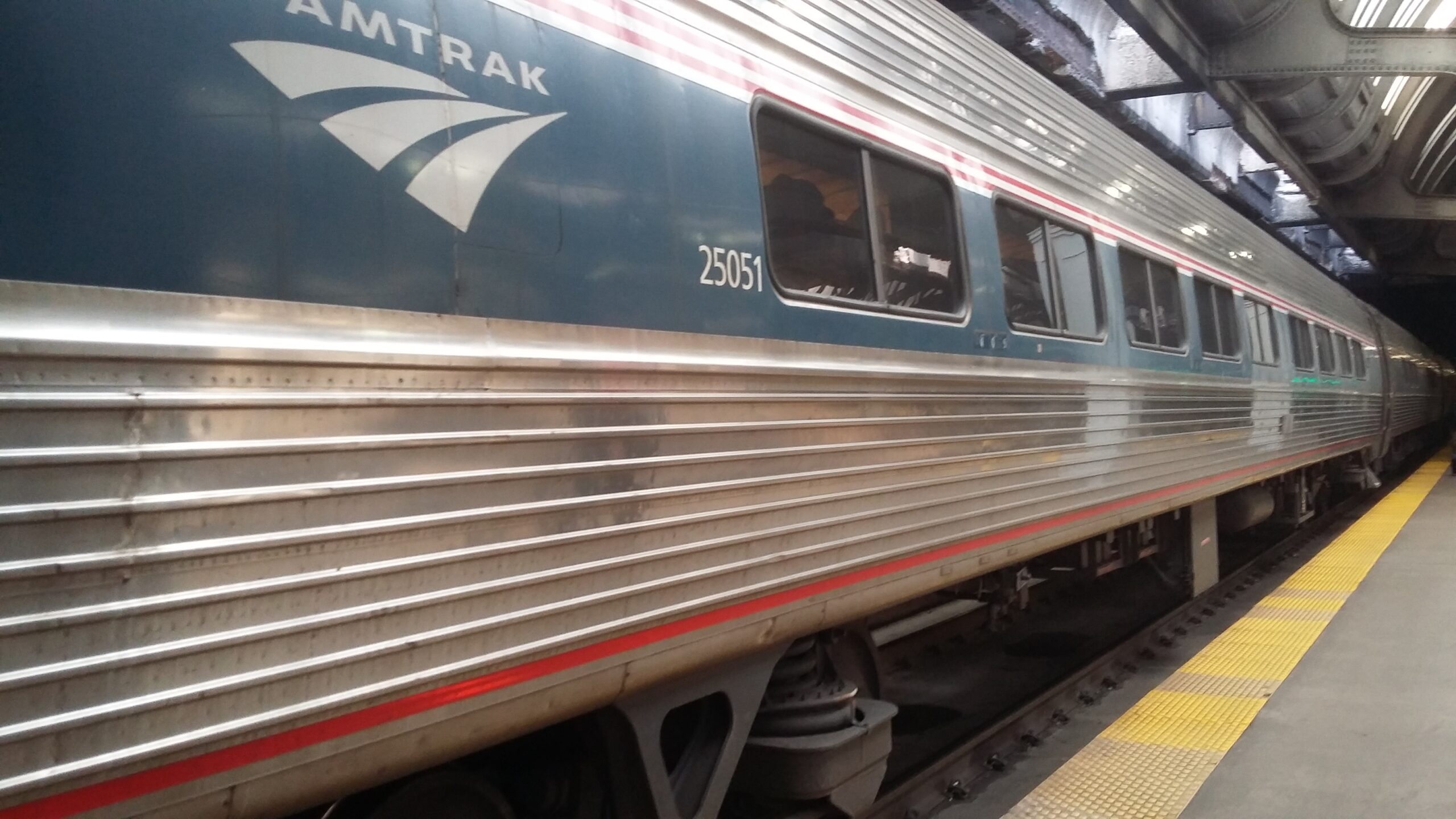 An image of an Amtrak car with the Amtrak logo in the foreground. It extends off into the distance toward the right side of the image. It is a passenger car with a wide blue stripe running along at window height and the rest of the body is polished metal. A thin red stripe also runs along the bottom and right above the blue stripe.