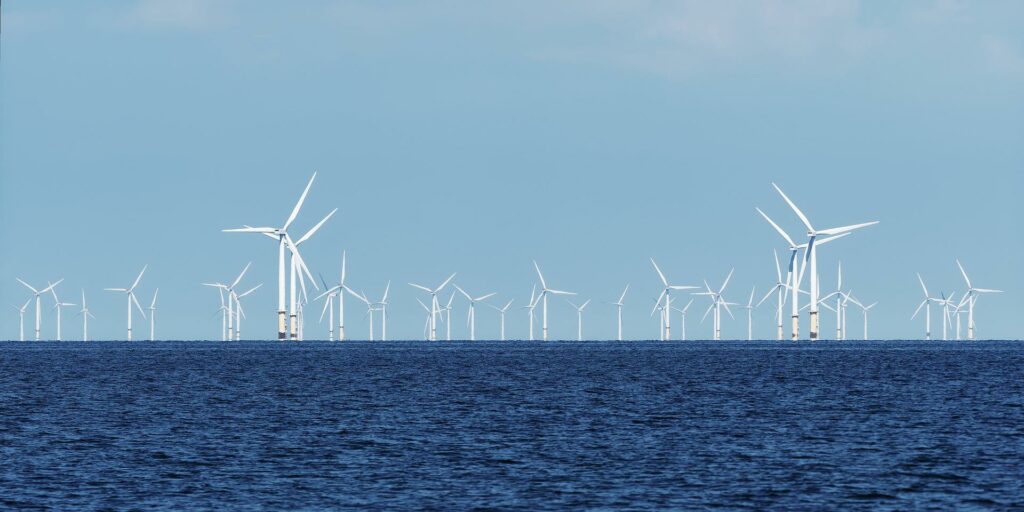 A large wind farm consisting of many large, three-bladed wind turbines sits in the ocean. The waves are a choppy dark blue and black while the sky is a light blue.