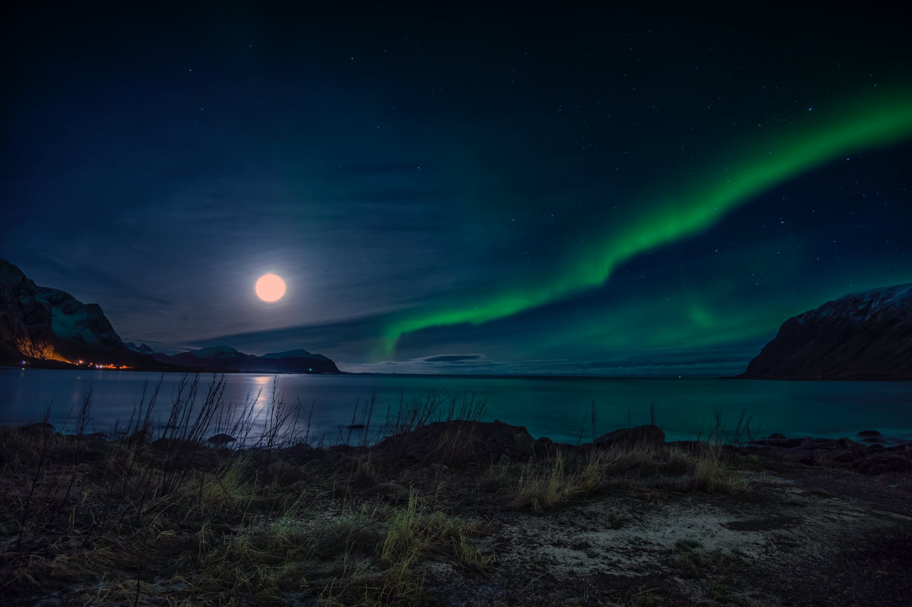 green aurora lights on the right hand side of the image are opposite a full moon on the left. The foreground is a sandy beach with some scrubby brush and what looks to be a town glows orange at the base of a mountain across the water.