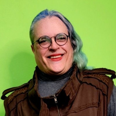 Photo of the author, a genderfluid individual with grey, black, and purple shoulder-length hair. They are smiling and wearing round black glasses and a brown zippered jacked with a heather grey turtleneck underneath.