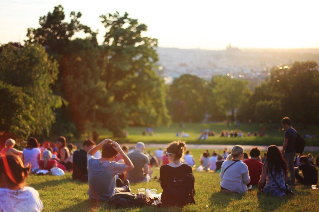 A number of people sitting in groups on green grass. There are green trees and a small mountain or hill in the background.