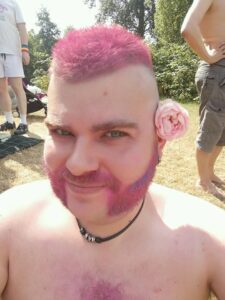 Image of a white male with pink hair and mutton chops. He has a pink flower over his left ear and a black necklace with three silver beads on it. There are trees and various other humans in the background.