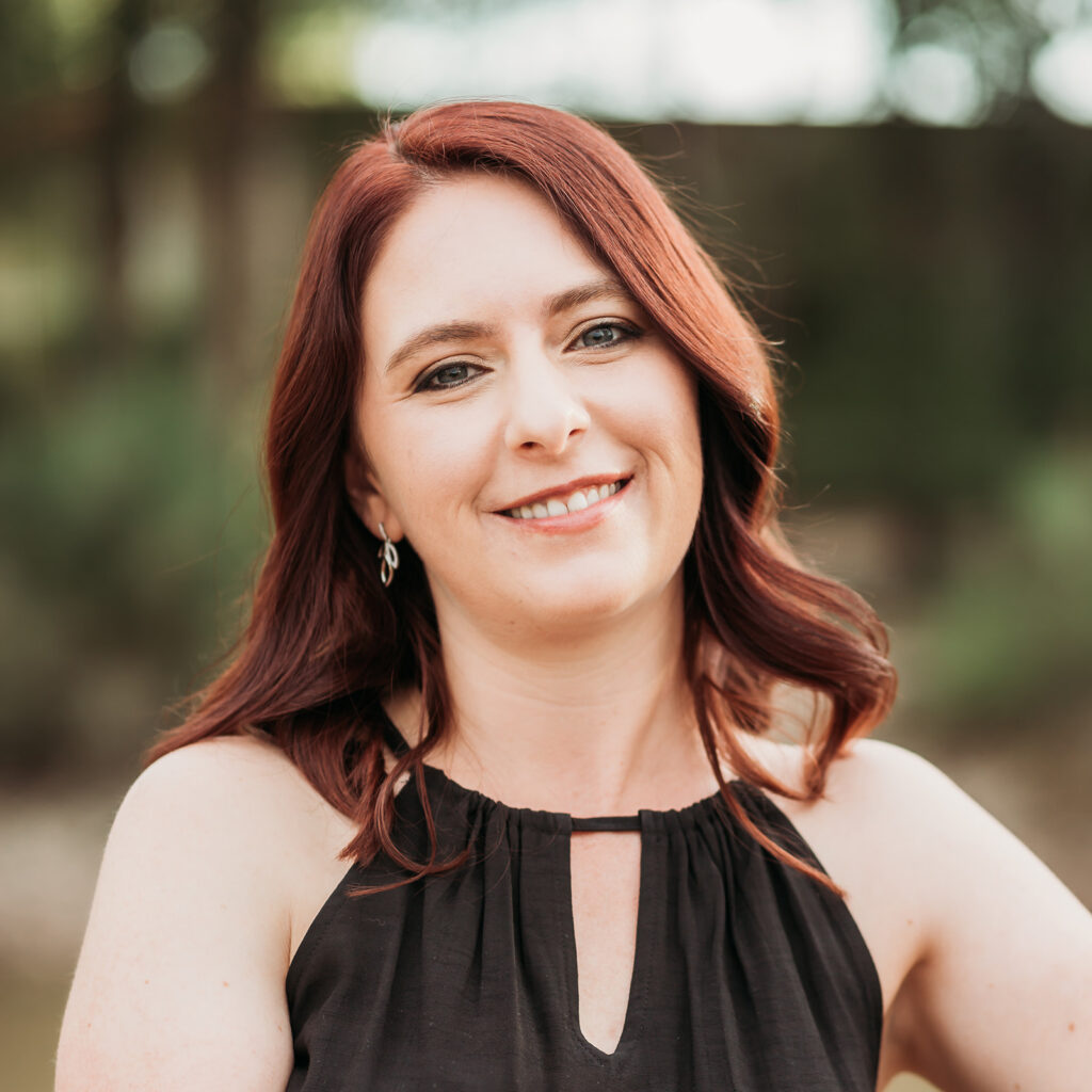 A woman with red hair wearing a black top with a keyhole. She is smiling and wearing small, silvery ear rings. 