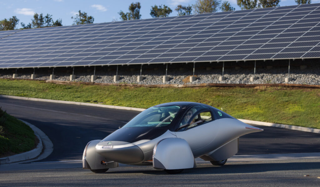 An aerodynamic three wheeled vehicle sits on blacktop in front of a solar array. The car has a white middle, silver nose, and black top and hood. The array is on a hill with grass at the bottom.